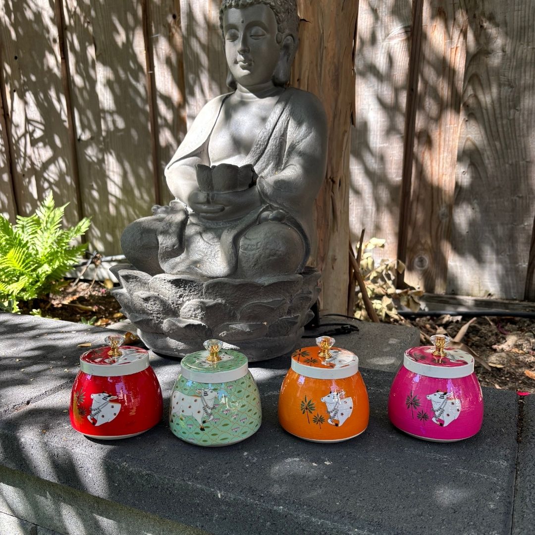 Four colorful indian pichwai jars in front of a stone Buddha statue outdoors.