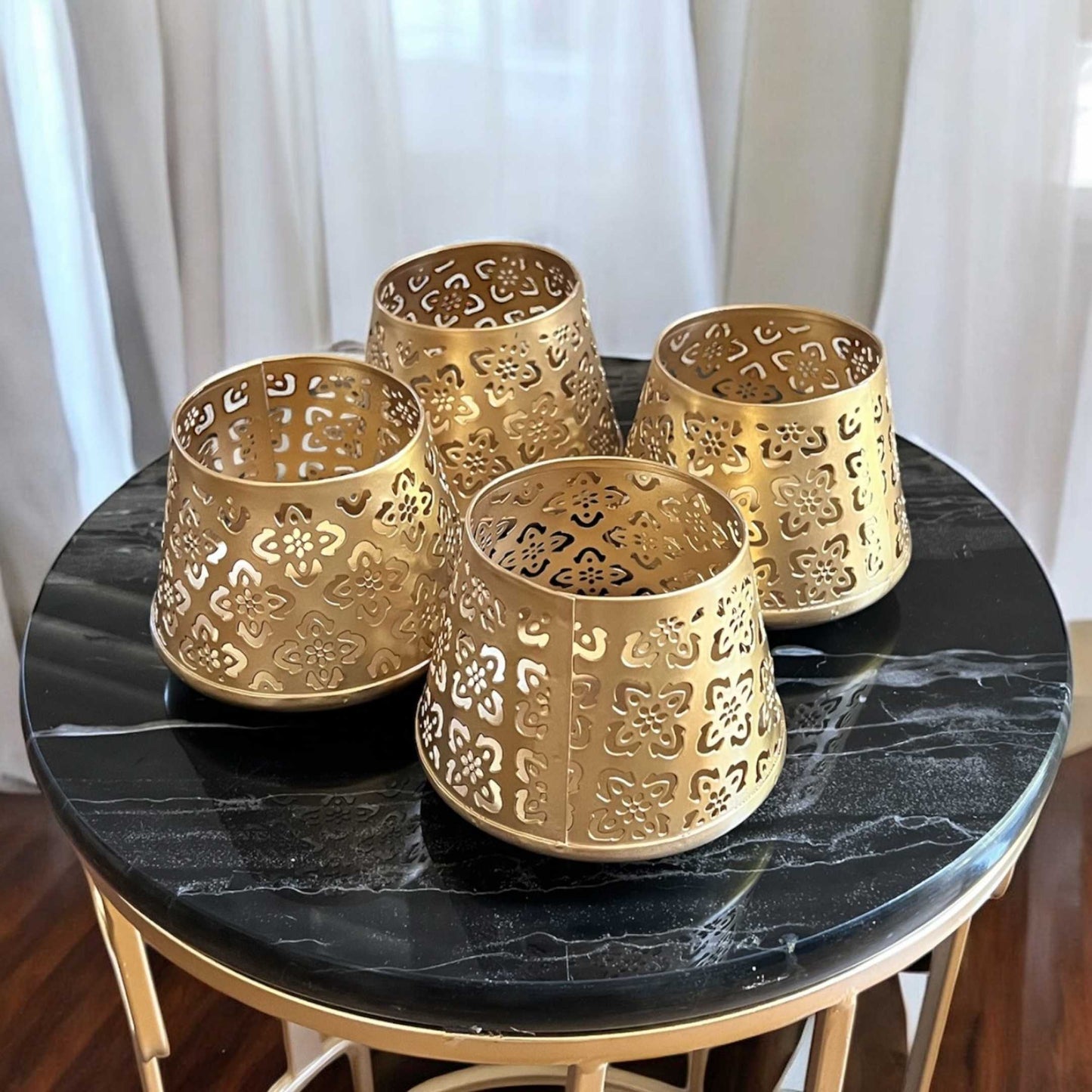 Four gold decorative candle holders on a black marble table with white curtains in the background.