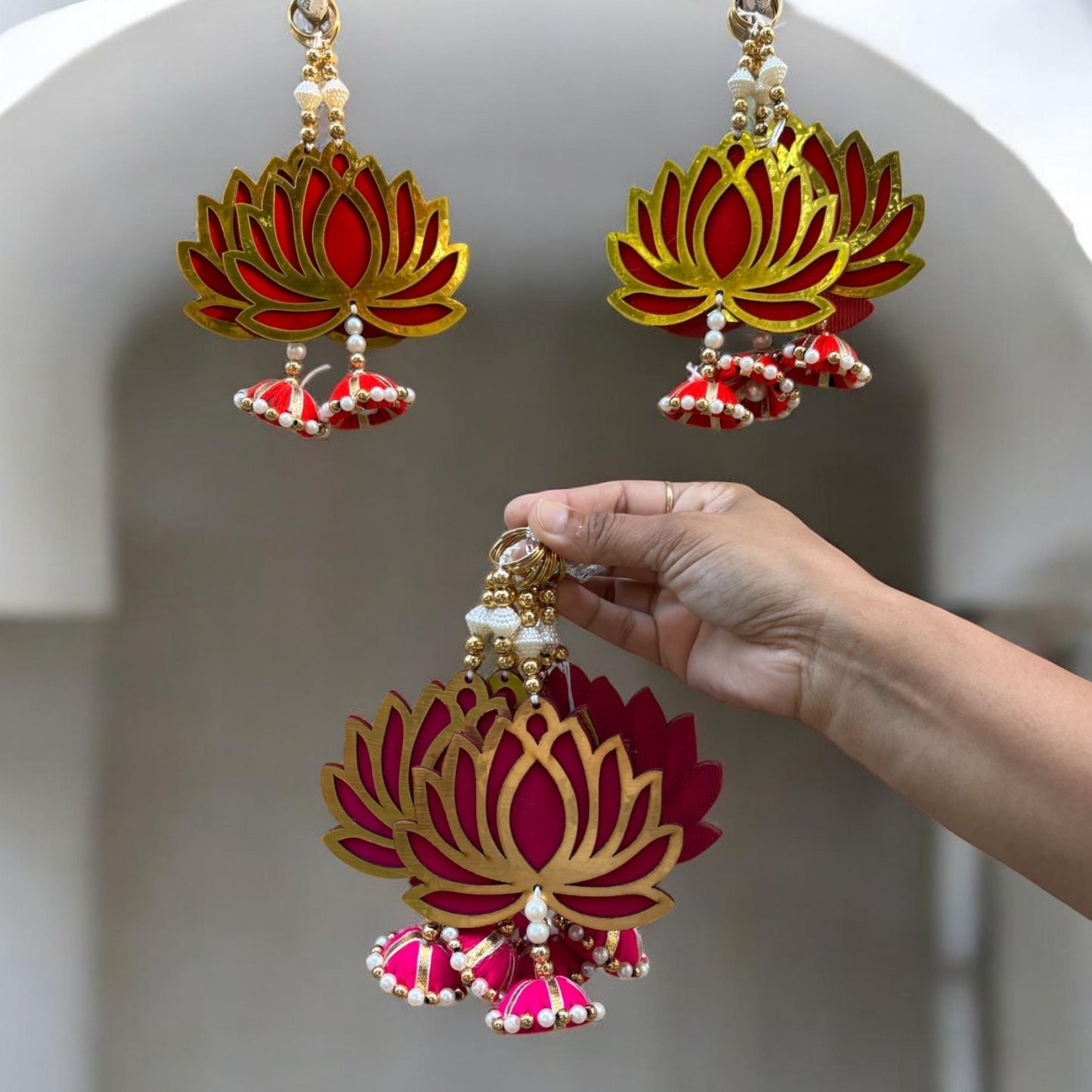 Lotus-shaped danglers with red, gold, and white colors held by a hand against a neutral background.