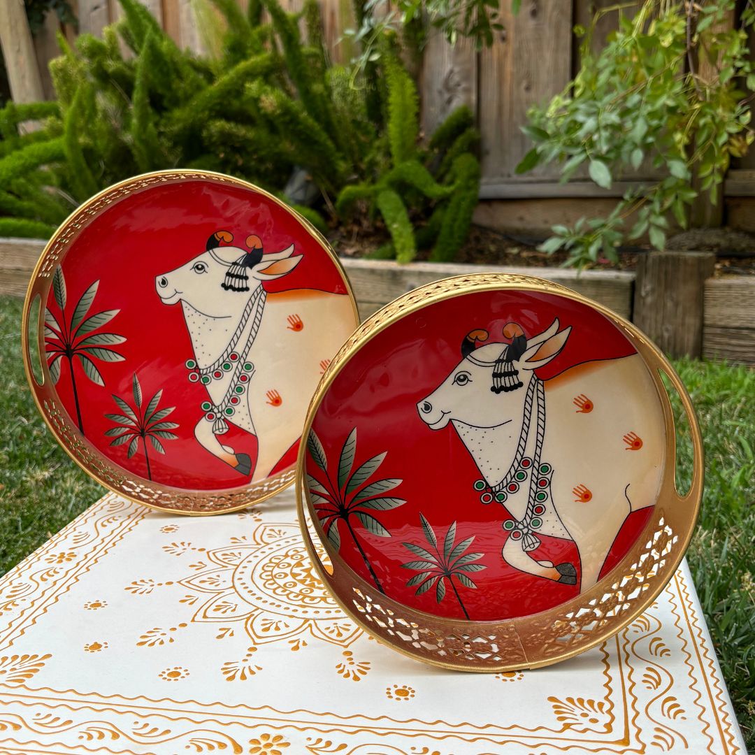 Two circular red metal trays painted with a white Pichwai-style cow motif, displayed upright against greenery to showcase design and color contrast.