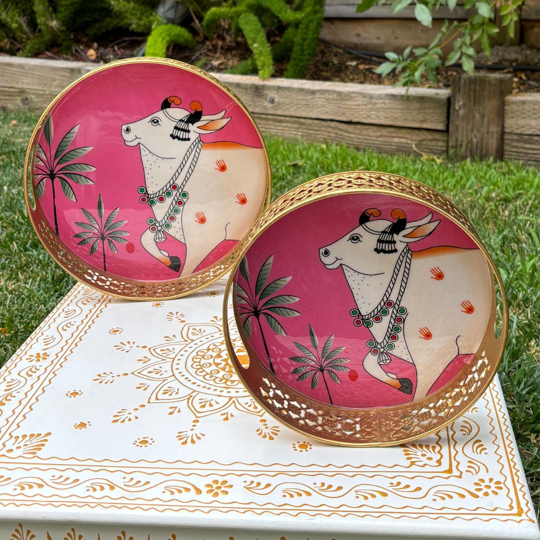 Two circular pink metal trays painted with a white Pichwai-style cow motif, displayed upright against greenery to showcase design and color contrast.