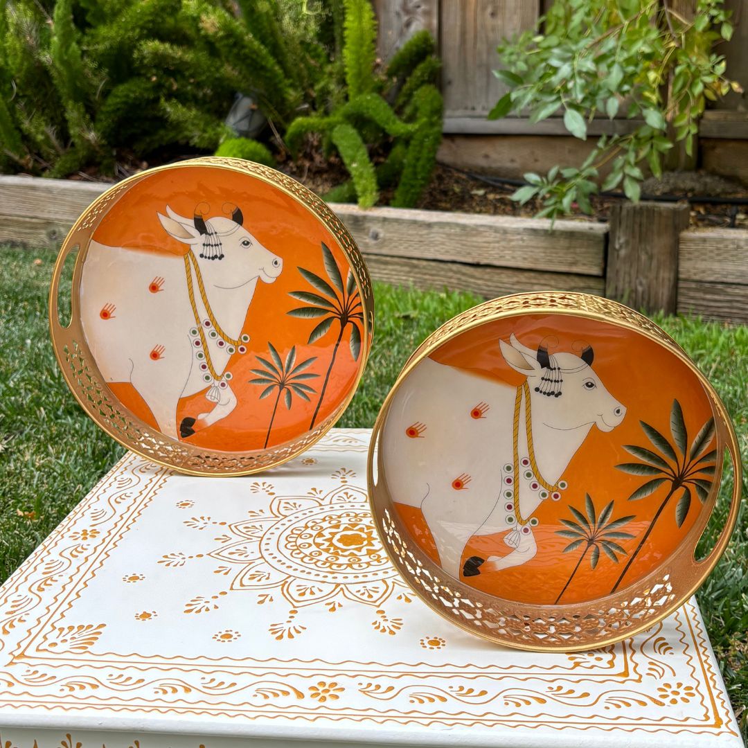 Two circular orange metal trays painted with a white Pichwai-style cow motif, displayed upright against greenery to showcase design and color contrast.