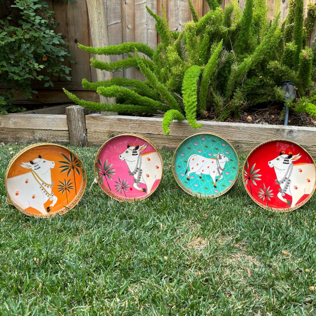 Four round metal trays in orange, pink, teal, and red, each featuring a white cow graphic in the center, arranged on grass to highlight variety and visual appeal.