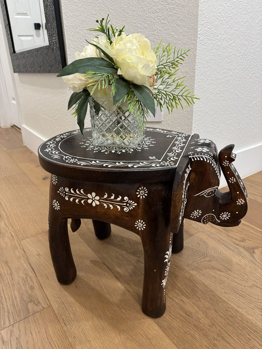 Dark brown carved mango wood elephant stool with white painted floral detail, holding a glass vase of white flowers in a room corner