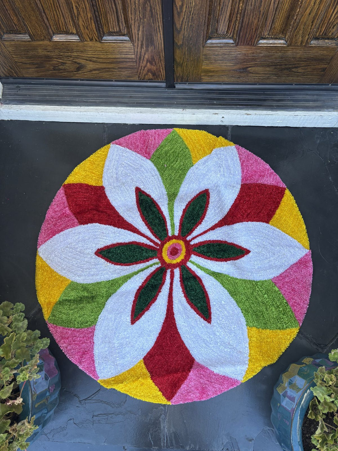 Round rangoli mat in white, pink, green and yellow wool yarn laid on stone at a dark wooden front door