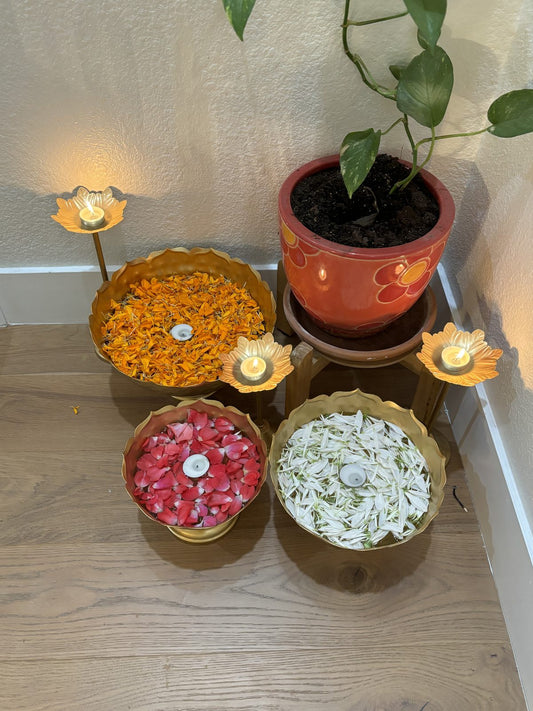 Two gold urli bowls filled with pink rose petals and white jasmine, with lit lotus tea lights alongside