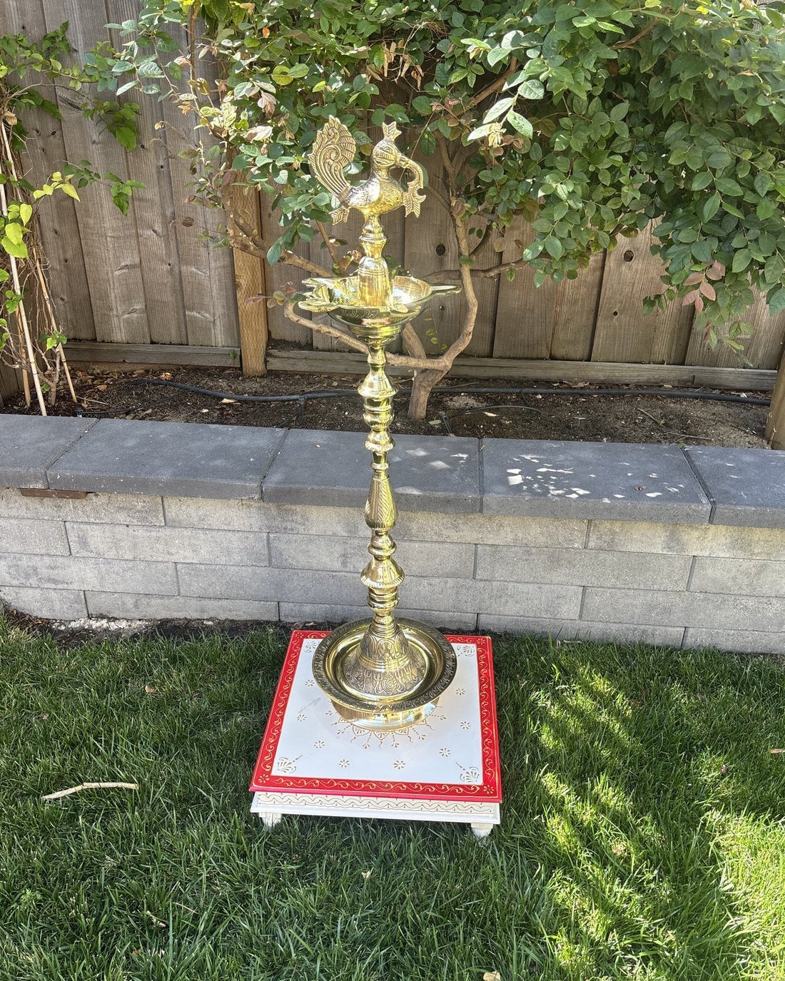 A tall brass nilavilakku oil lamp standing at full height on a white and red painted wooden chowki