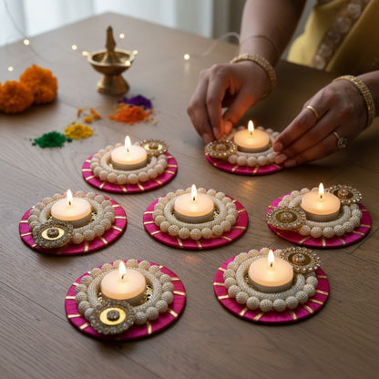Seven pink and pearl beaded t-light holders with gold accents, lit and arranged on a table with a brass lamp and coloured rangoli powder