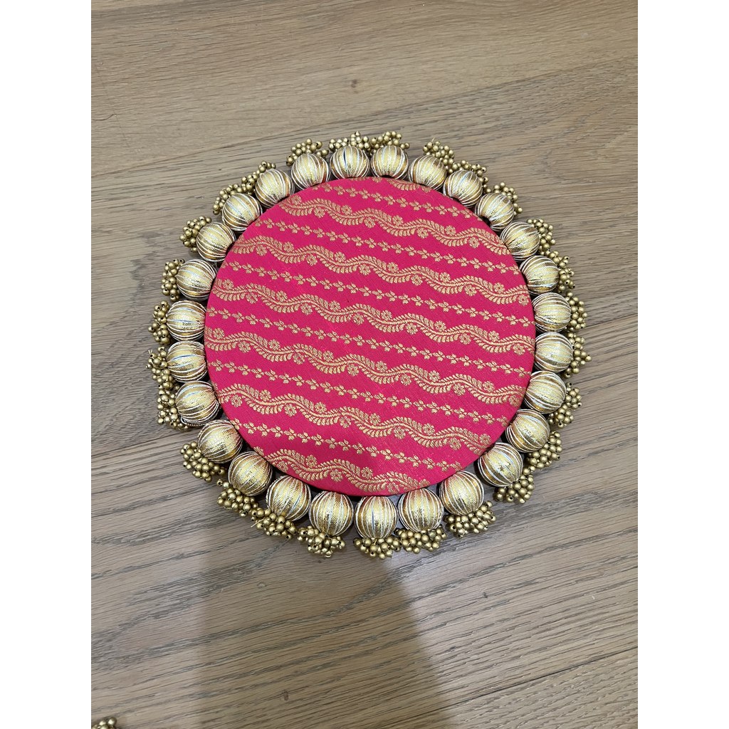 Single round Patola brocade mat in pink with gold wavy stripes and a gold beaded border, on a wooden floor.