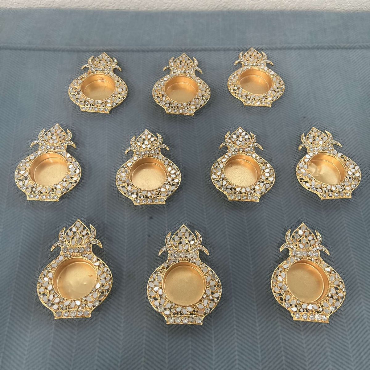 Ten gold mirror kalash tea light holders in rows on a blue herringbone cloth