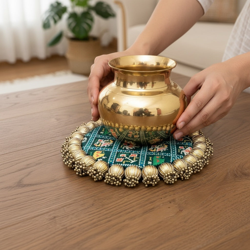 Hands placing a large engraved brass kalash onto a teal Patola brocade mat with a gold beaded border.