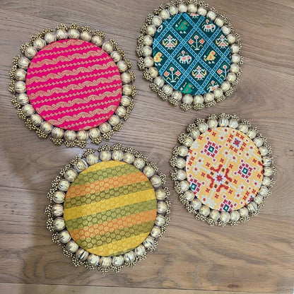 Set of four round Patola brocade mats on wood in pink and gold stripe, teal geometric, yellow and green stripe, and cream with red Patola.