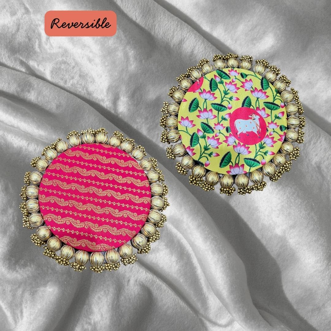 Two round Patola brocade mats on silver satin, one pink with gold wavy stripes and one yellow floral with a pink medallion.