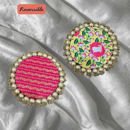 Two round Patola brocade mats on silver satin, one pink with gold wavy stripes and one yellow floral with a pink medallion.