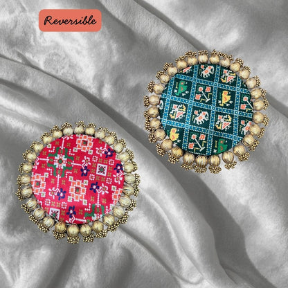 Two round Patola brocade mats on silver satin, one red with Patola geometry and one teal with a grid motif, showing the reversible prints.