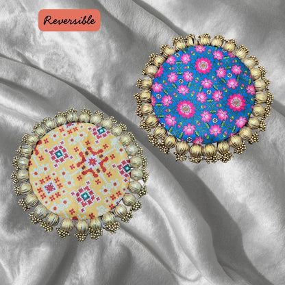 Two round Patola brocade mats on silver satin, one cream with red Patola geometry and one blue scattered with pink flowers.