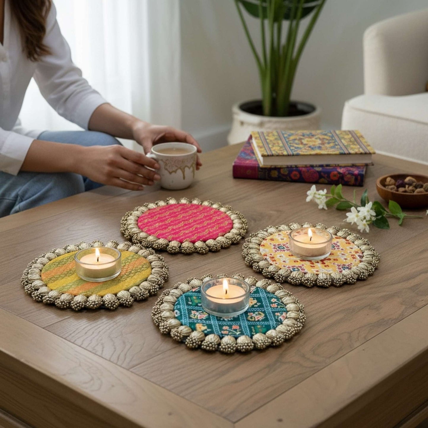 Four round Patola brocade mats with gold beaded borders on a wooden coffee table, three holding lit tealights beside a seated person with a mug.