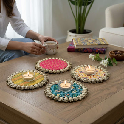 Four round Patola brocade mats with gold beaded borders on a wooden coffee table, three holding lit tealights beside a seated person with a mug.