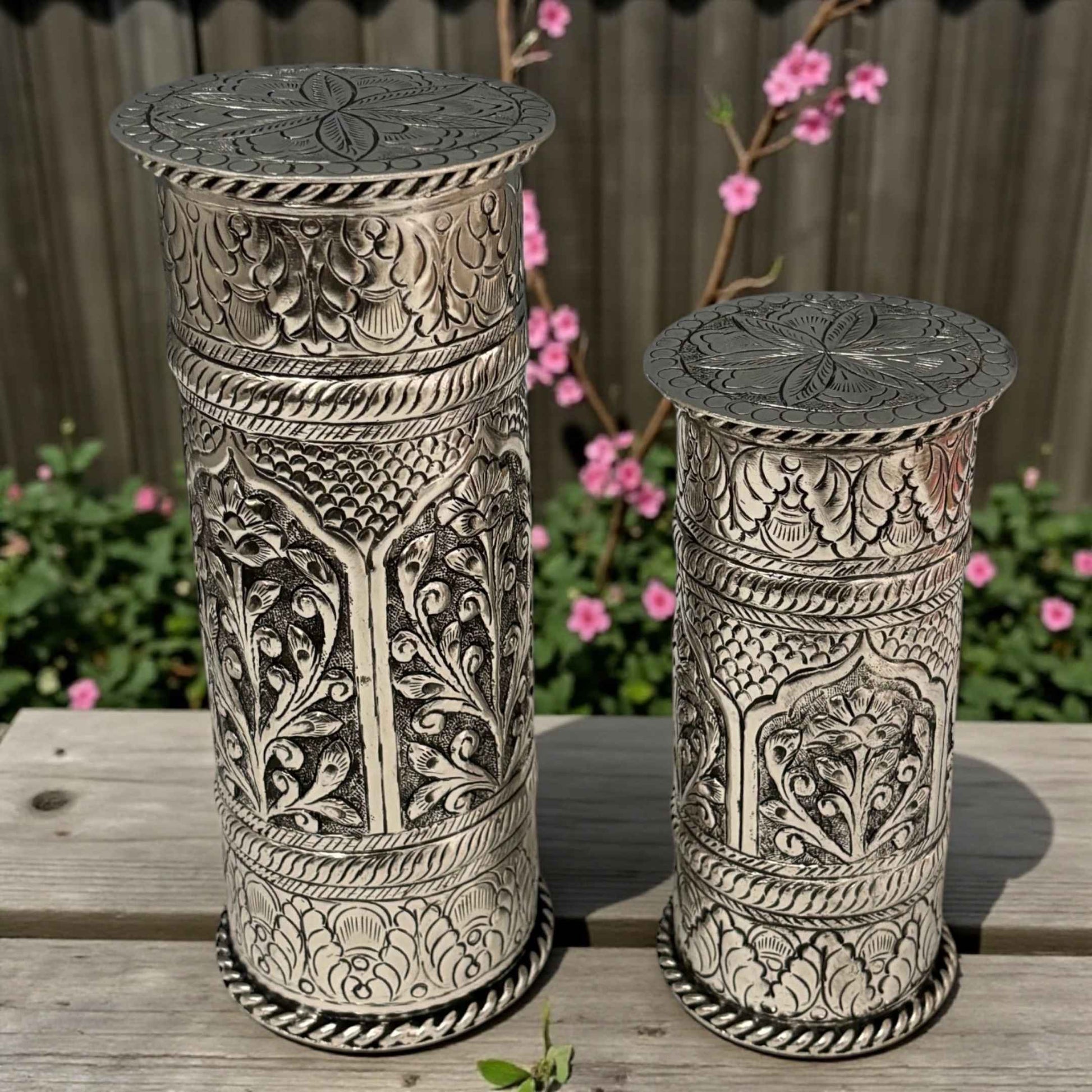 Two ornate cylindrical metal stands with intricate designs on a wooden surface.