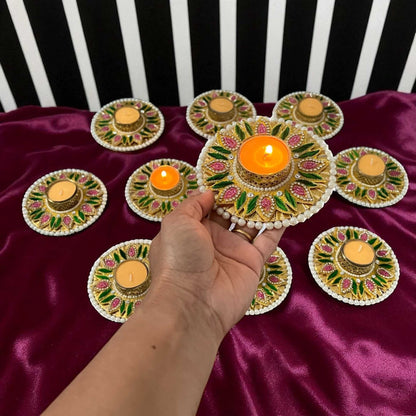 Hand holding lit Diwali diya; set of 10 T-light holders on satin backdrop with pearl rim and floral detailing.