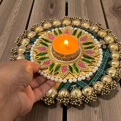 Lit tealight diya placed on festive patola mat; multicolor pearl and stone work holder held by hand — traditional Indian decor.
