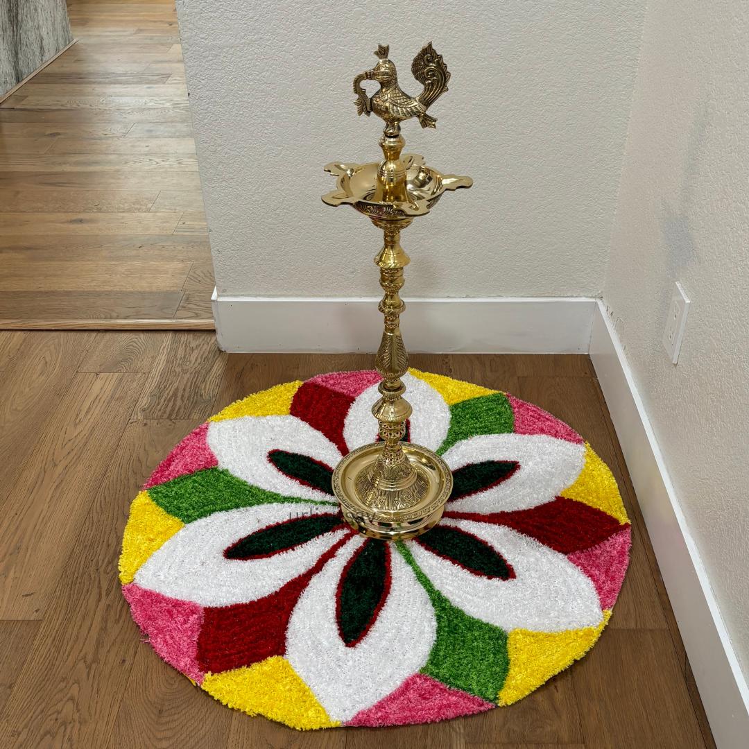 Round rangoli mat with a white flower design in pink, yellow, and red segments, brass diya lamp standing at center