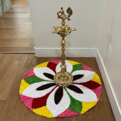 Round rangoli mat with a white flower design in pink, yellow, and red segments, brass diya lamp standing at center