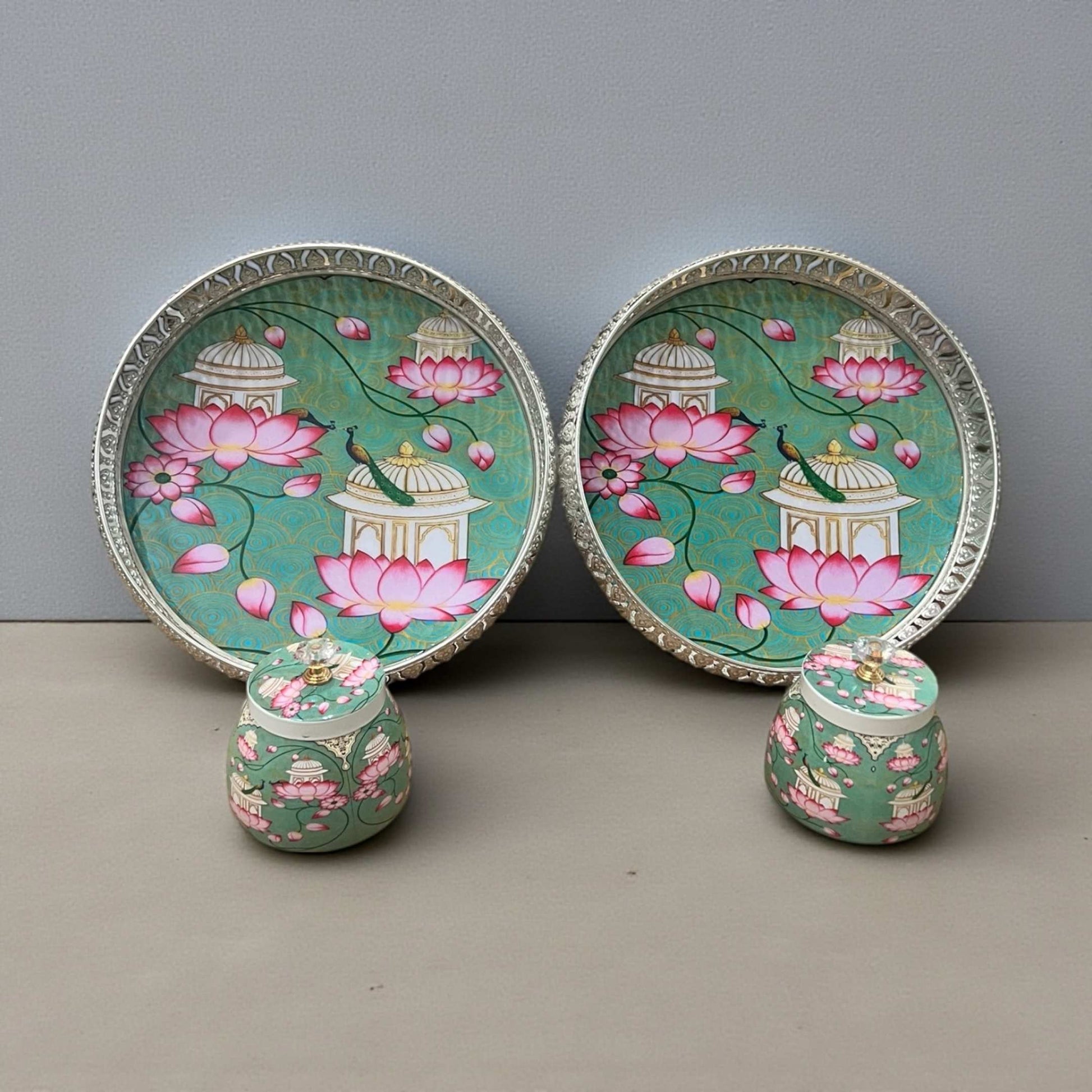 Two decorative German silver Pichwai art trays with two matching storage jars on gray background