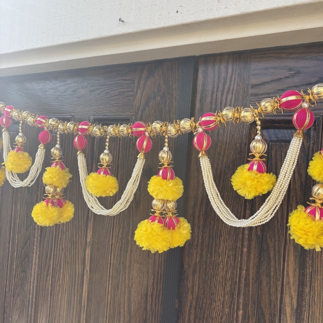Pink and gold pearl toran bandhanwar with yellow marigold pom-poms on dark wood door — handmade Indian Bandhanwar door garland