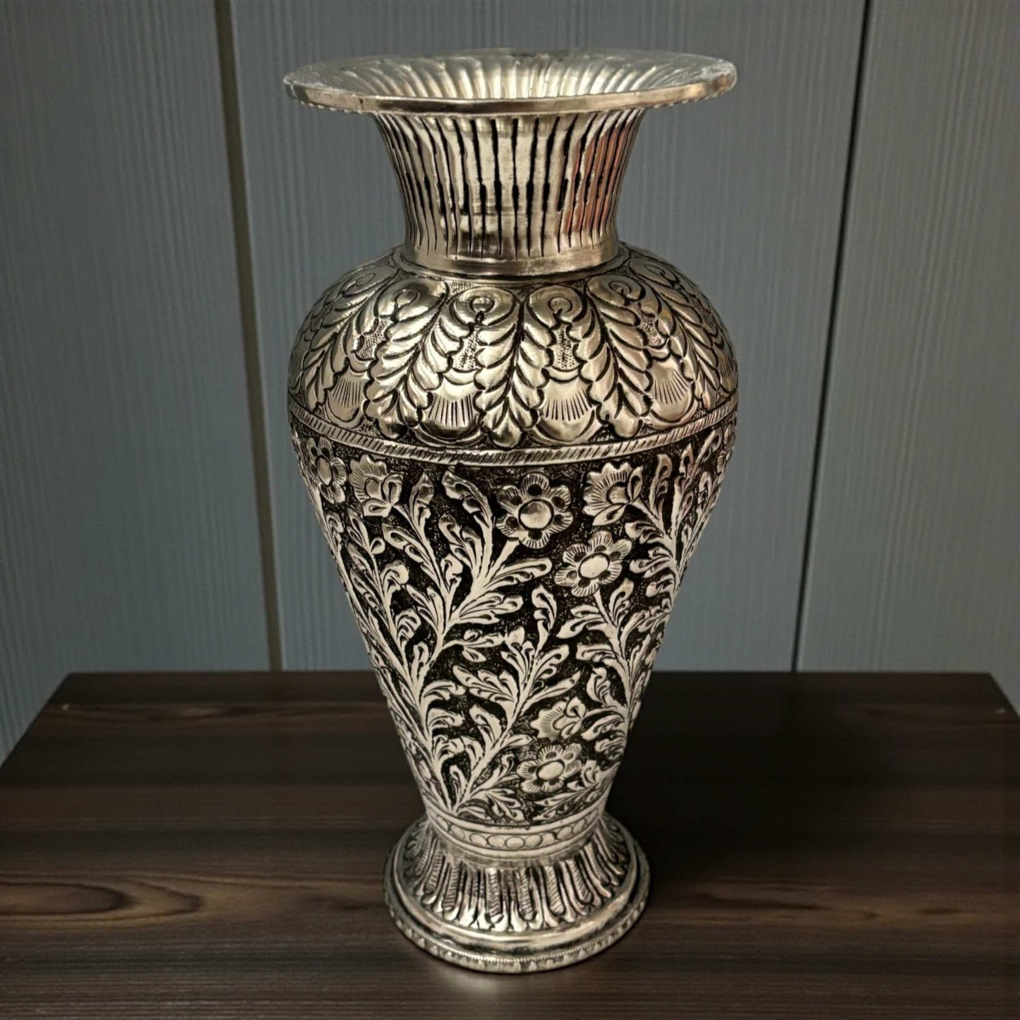A sophisticated tall silver vase showcasing fine vine etchings against a dark, elegant paneled background.