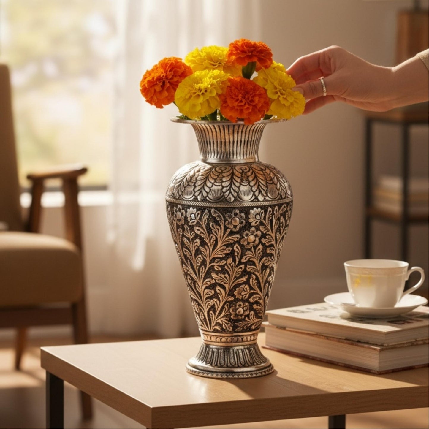 A hand arranging vibrant yellow and orange marigolds in a silver-plated copper vase on a sunlit table.