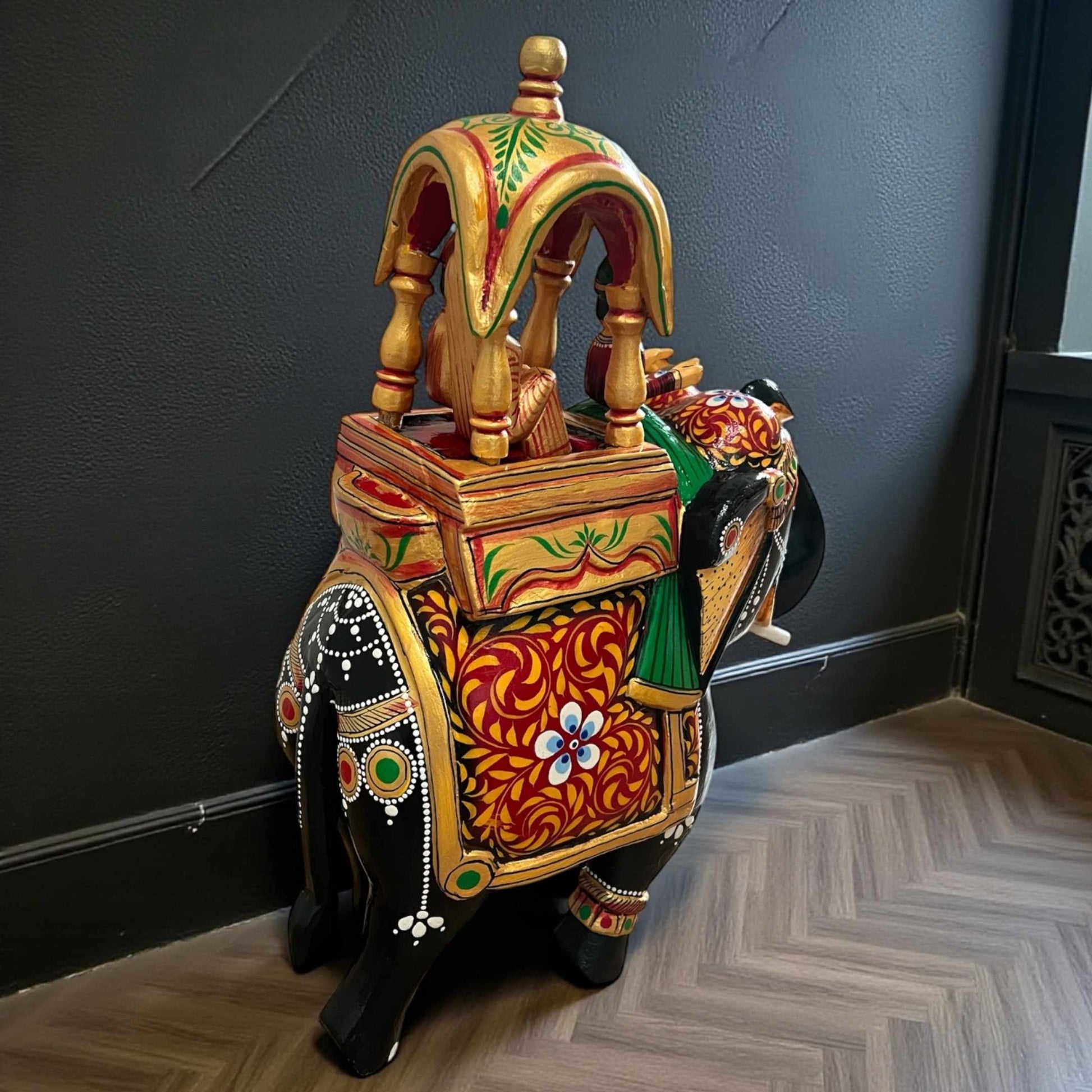 Walking Ambabari Elephant handcrafted wooden statue with ornate carved detailing positioned against dark textured wall for dramatic contrast.