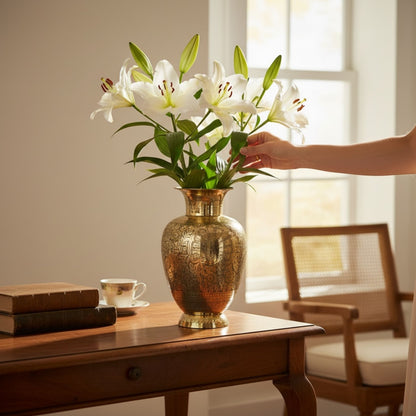 Hand arranging white lilies in a gold vase on a wooden table with a chair and window in the background.