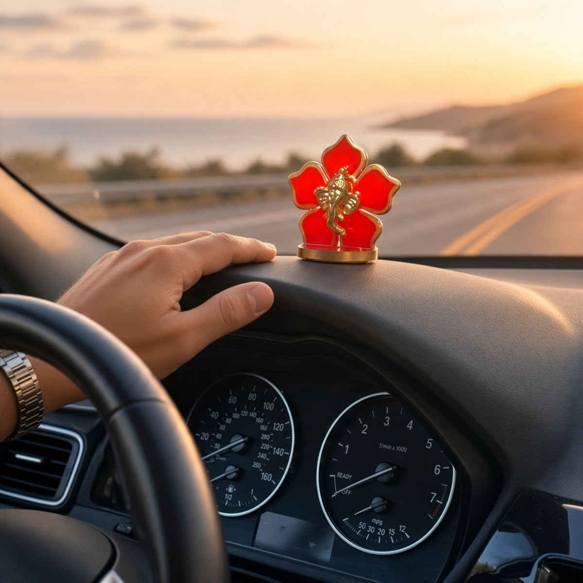 Red flower Ganesha car idol on a car dashboard as a driver steers along a coastal road at sunset