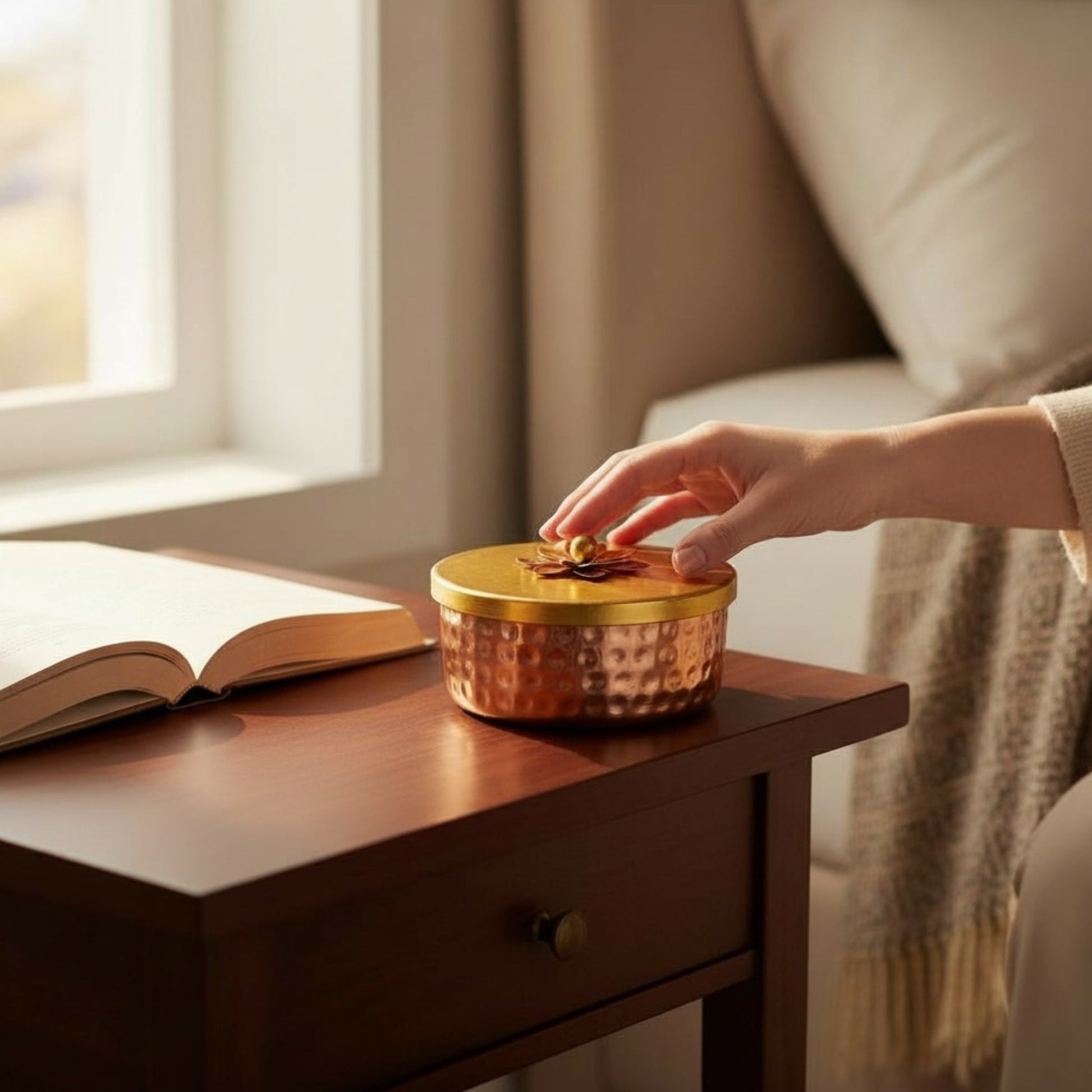 Hand opening the lid of a small copper container beside a book — showing lid design and usability