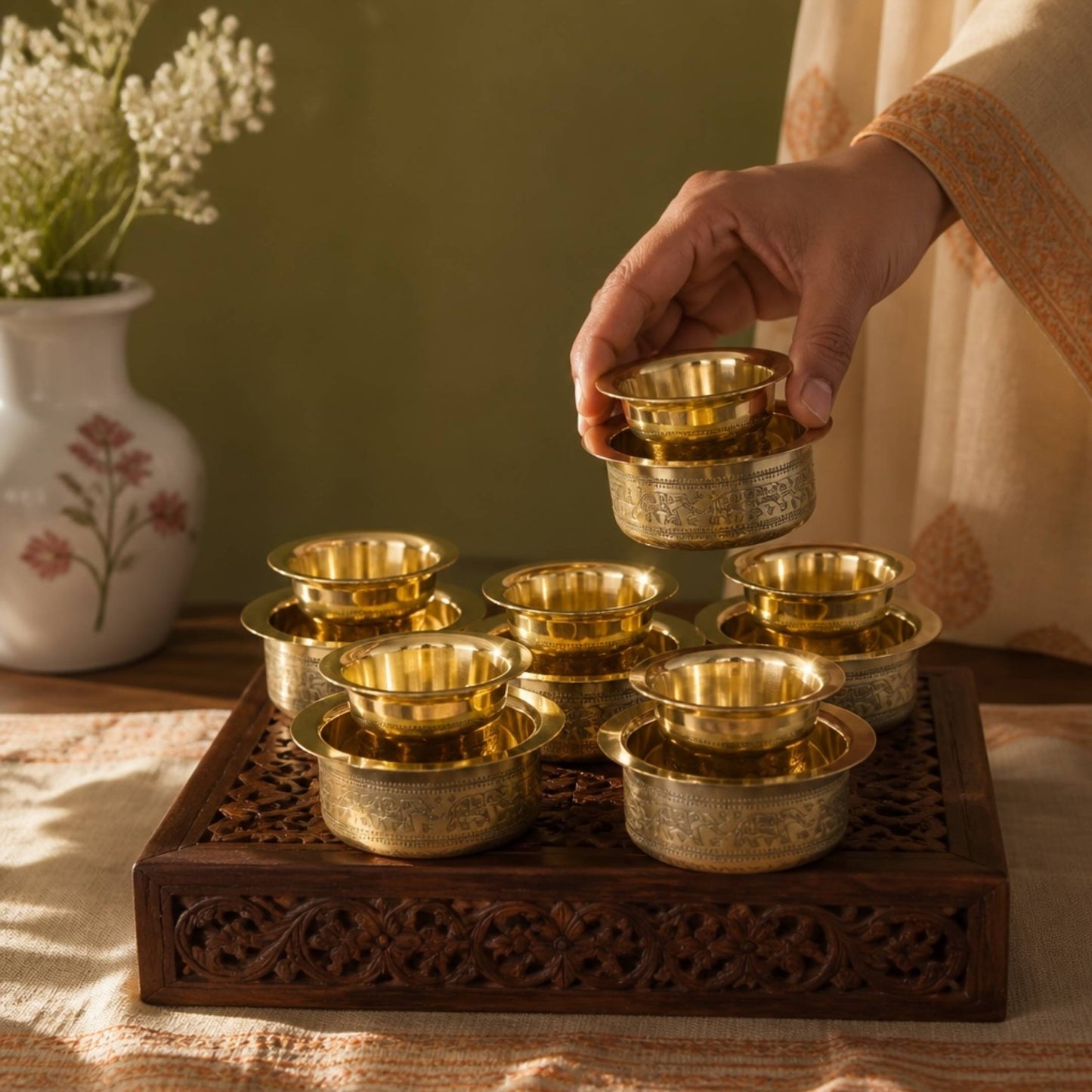 Hand lifting one handmade brass davara bowl from carved wooden tray with five matching pieces