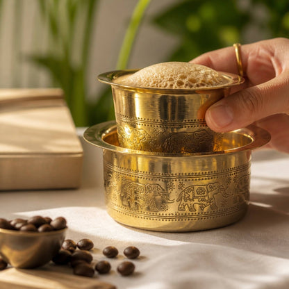 Hand holding handmade brass davara tumbler with frothy South Indian filter kaapi and coffee beans