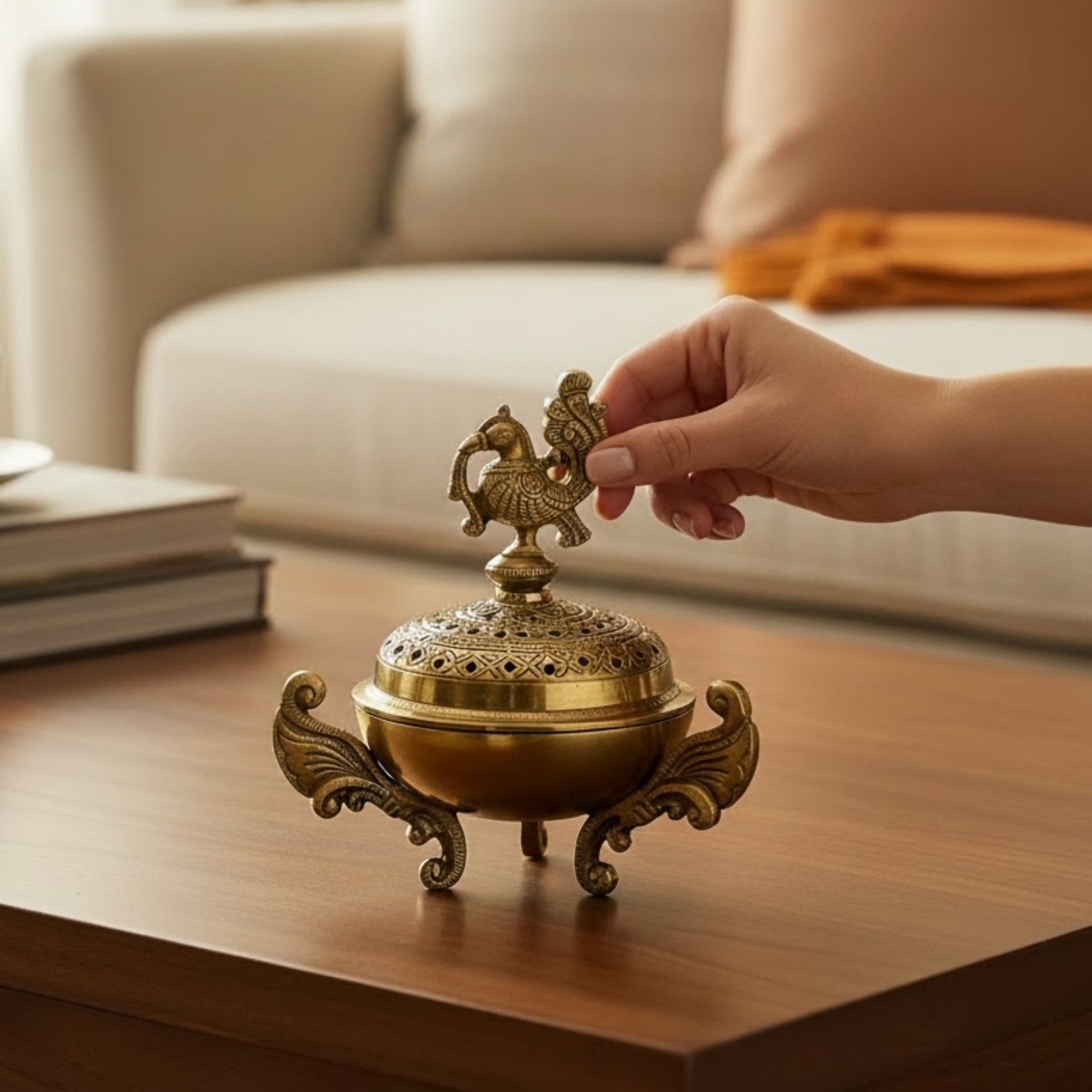 Brass dhoop dani on a wood coffee table with a hand lifting the peacock finial lid near books