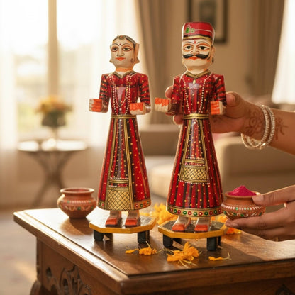 Red hand-painted Isar Gangaur King and Queen dolls held above a wood table with a kumkum powder pot and small clay pot