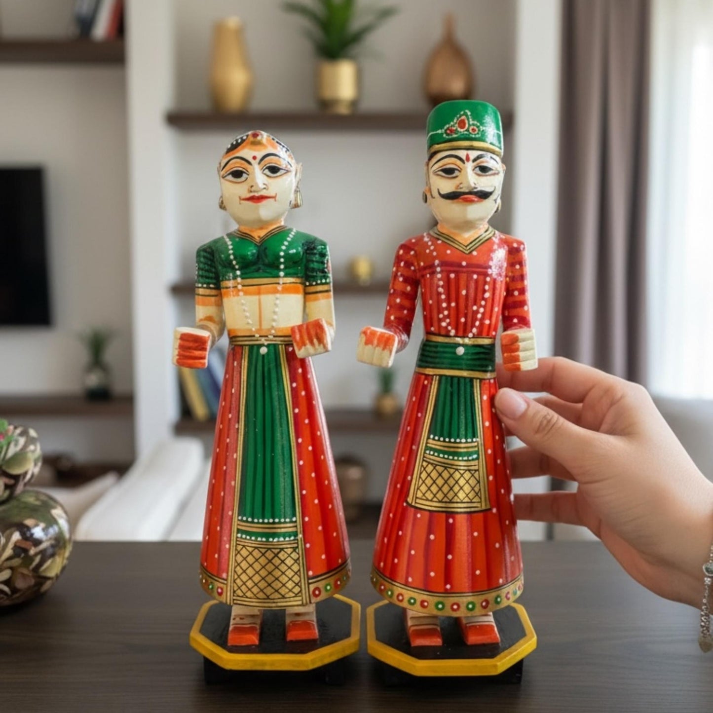 Green and red hand-painted Isar Gangaur King and Queen dolls displayed on a wood console with vases and shelving behind