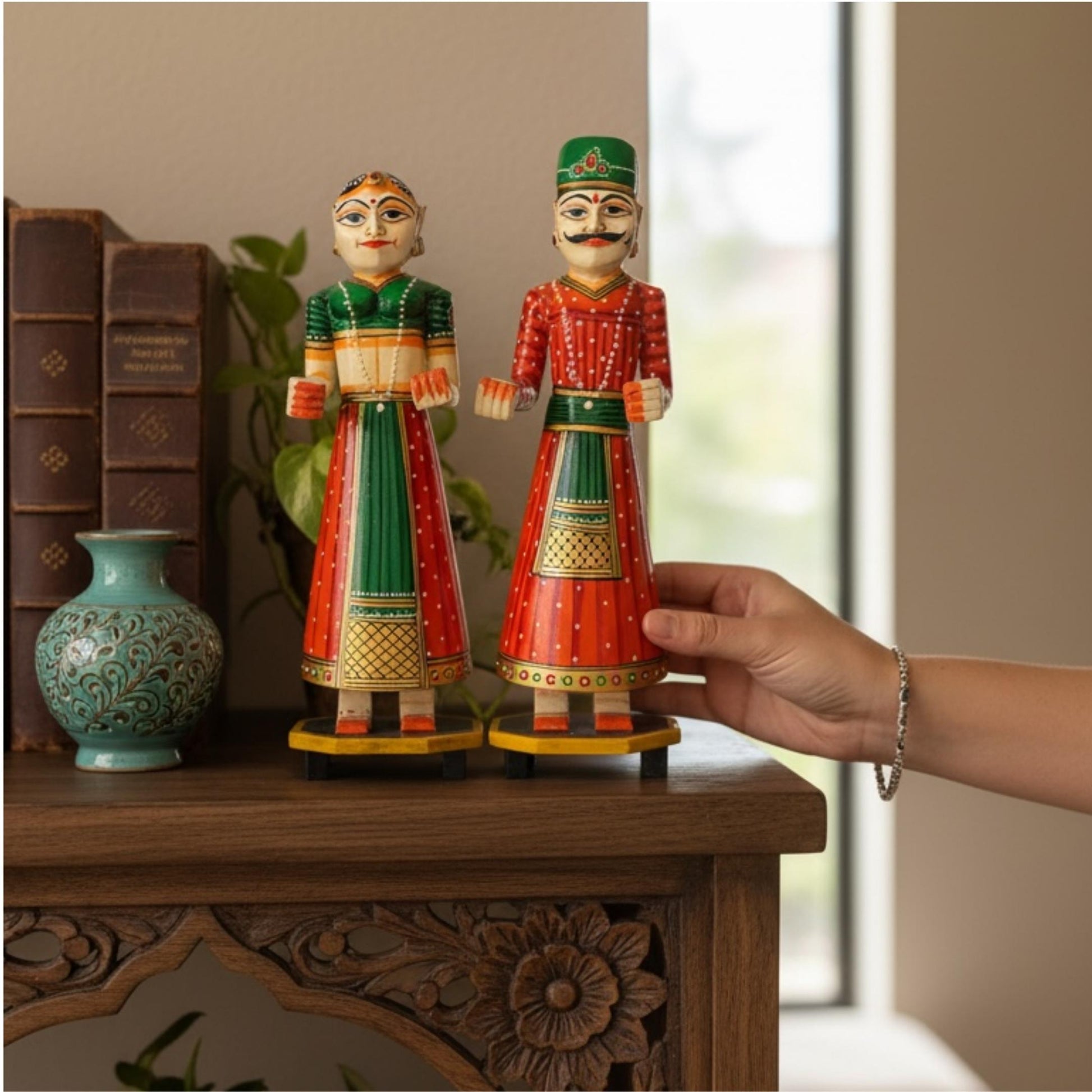 Green and red hand-painted Isar Gangaur King and Queen dolls displayed on a carved wood console beside a teal vase