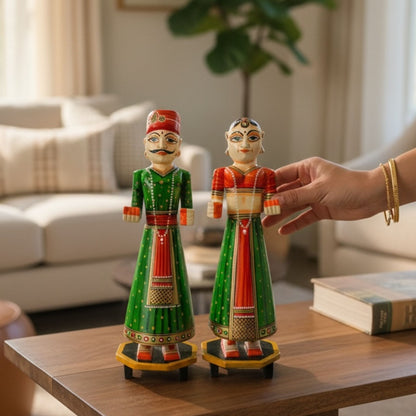Green and red hand-painted Isar Gangaur King and Queen dolls displayed on a wood table beside a potted plant and a book