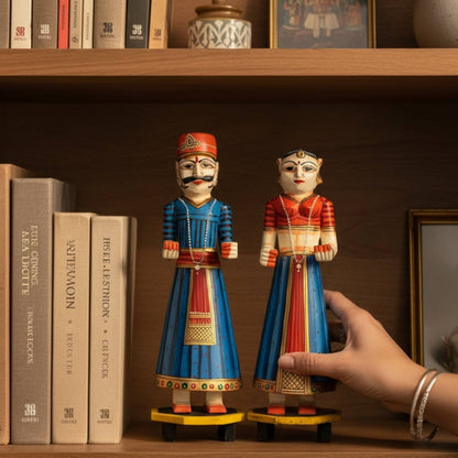 Blue and red hand-painted Isar Gangaur King and Queen dolls displayed on a bookshelf beside books and a framed photo