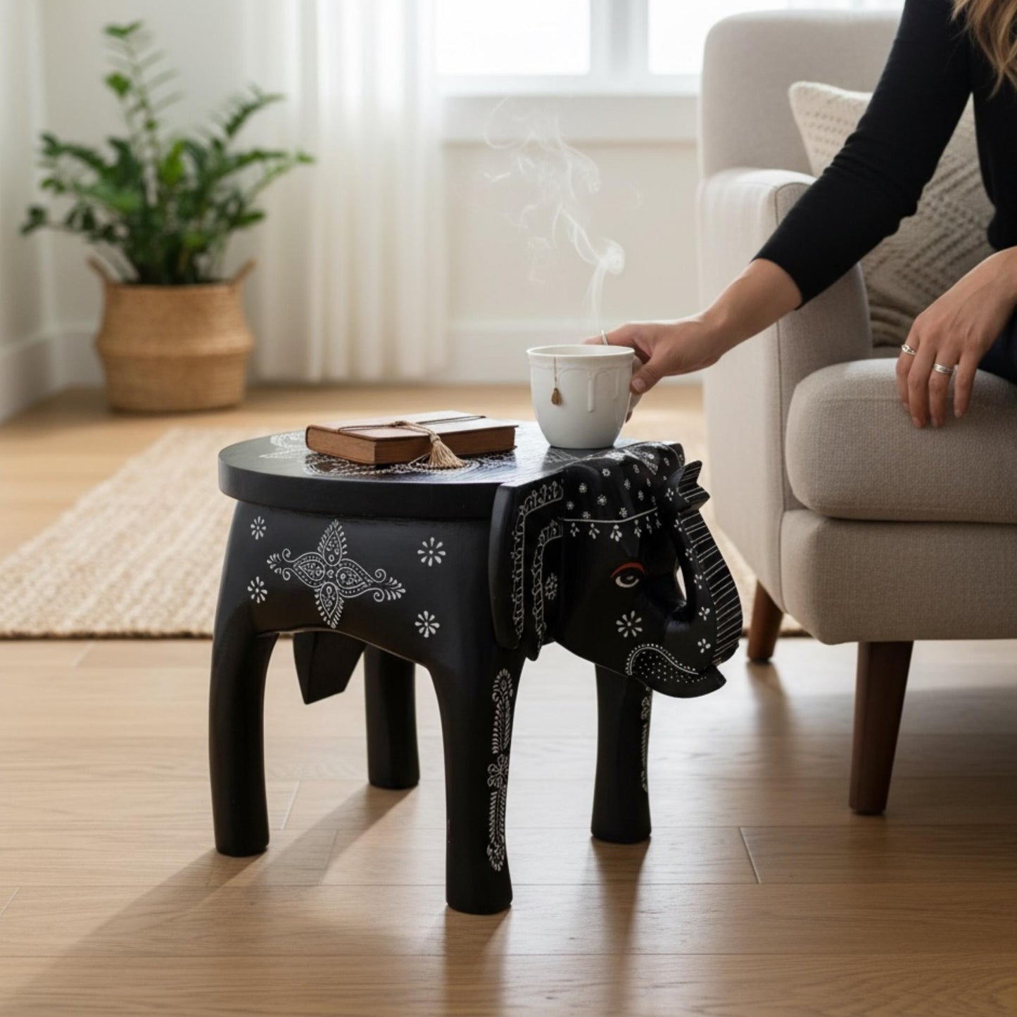 Black elephant stool with a white mandala top holds a steaming teacup and a book beside a living room couch