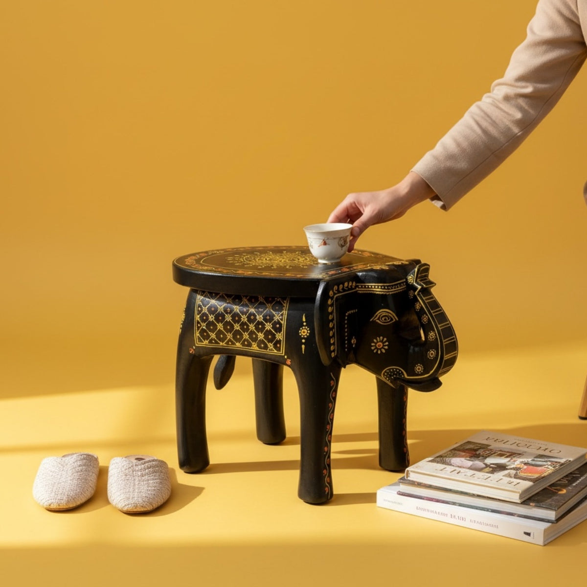 Black and gold elephant stool on a yellow backdrop with a hand placing a teacup, slippers, and books nearby