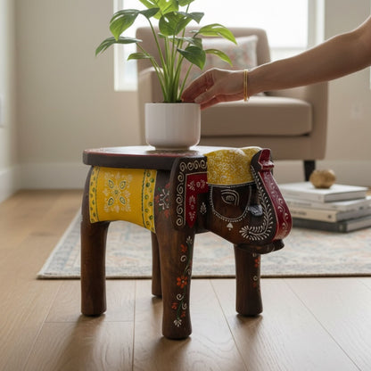 Elephant stool with a red and yellow painted top and saddle, hand placing a potted plant on top