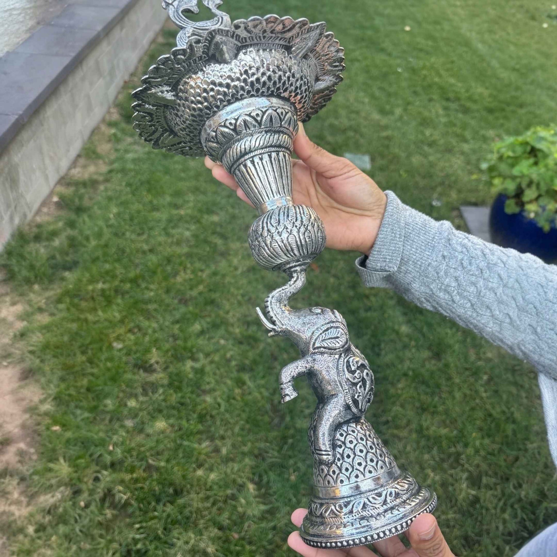 Person holding German silver peacock elephant base samai oil lamp outdoors – Urli Utsav decor