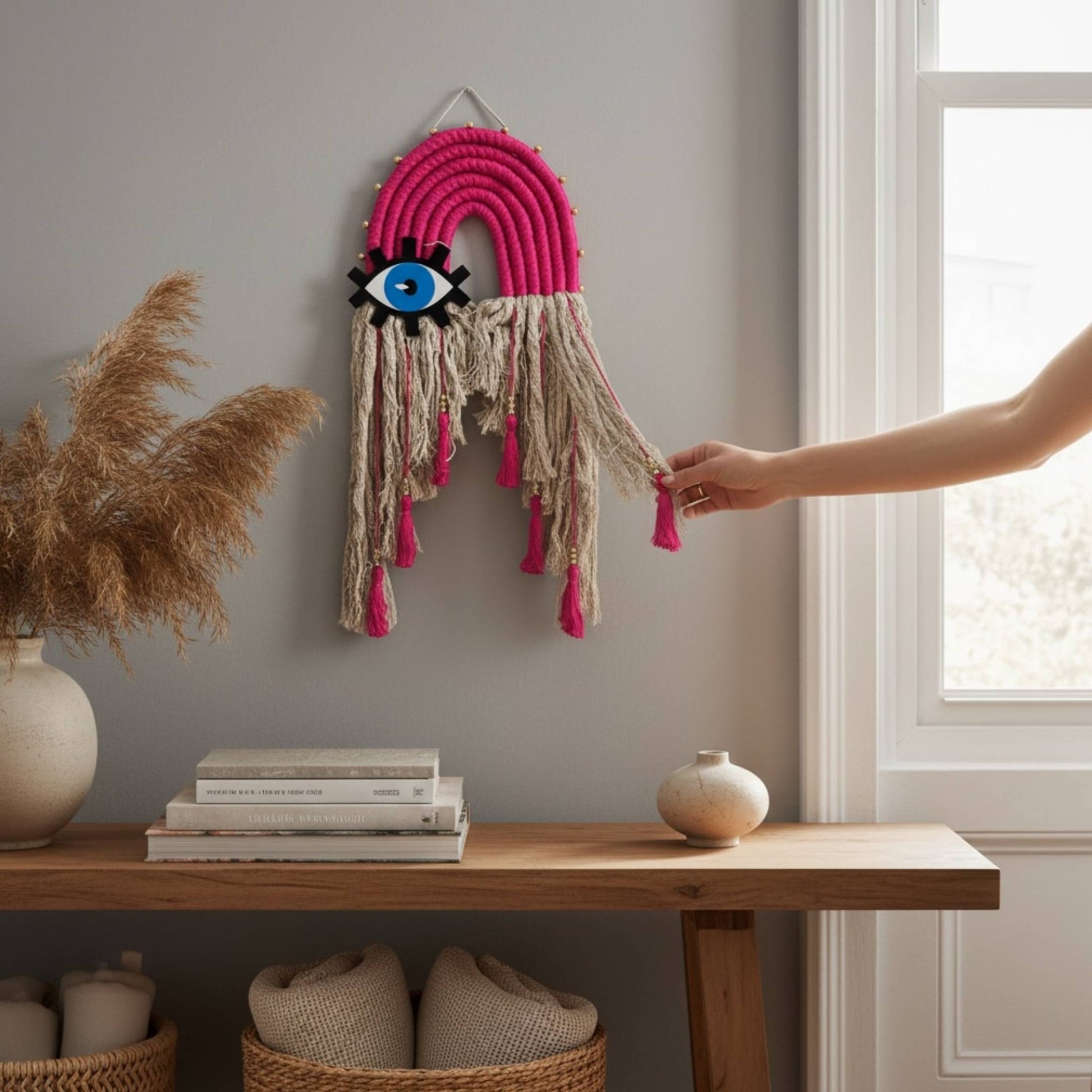 Bohemian style pink and beige strings and a blue eye design evil eye wall decor hanging above a modern table in a bright living room.