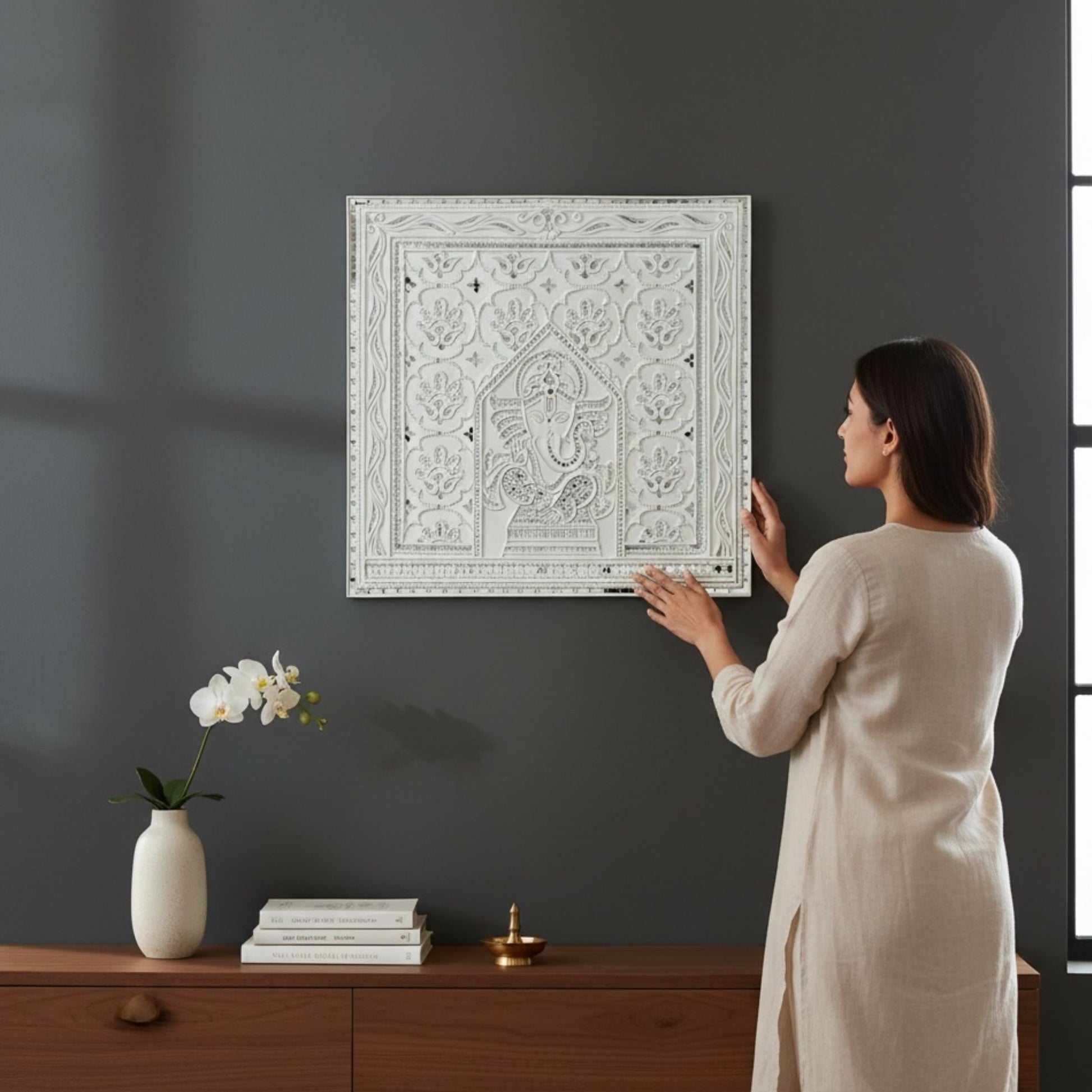 Woman hanging 2ft Ganesha White Lippan wall art piece on a dark gray wall.