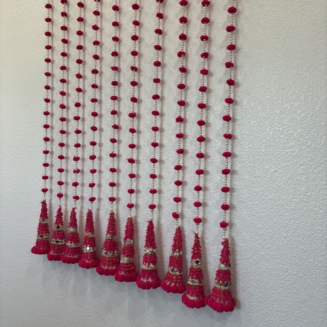 A decorative curtain made of multiple vertical strands of red pom-poms and tassel-like beads hanging against a plain off-white wall.